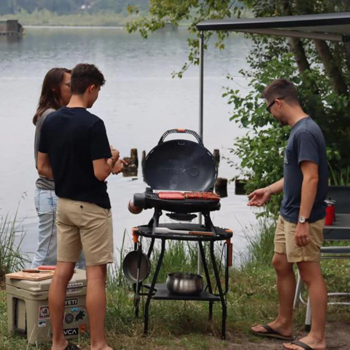 A black o-grill 500 being used to grill some burgers and kebabs beside a lake